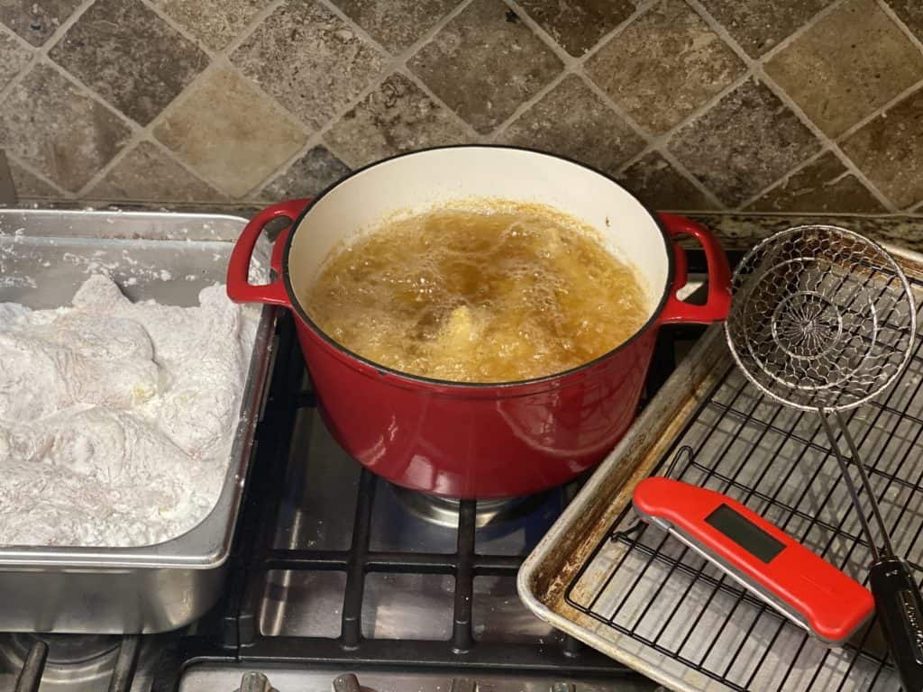 Chicken Legs Dredged in Potato Starch in a stainless container next to a red pot of bubbling hot oil and then a drying rack with a red thermometer and fry spider sitting on top