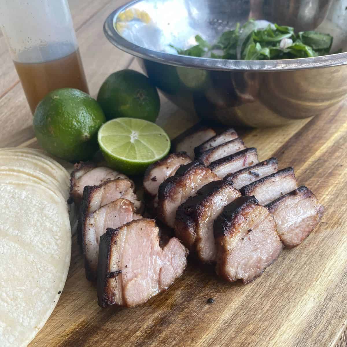 Sliced Pork Belly on a wood board with tortillas, lime, chipotle oil and a stainless steel mixing