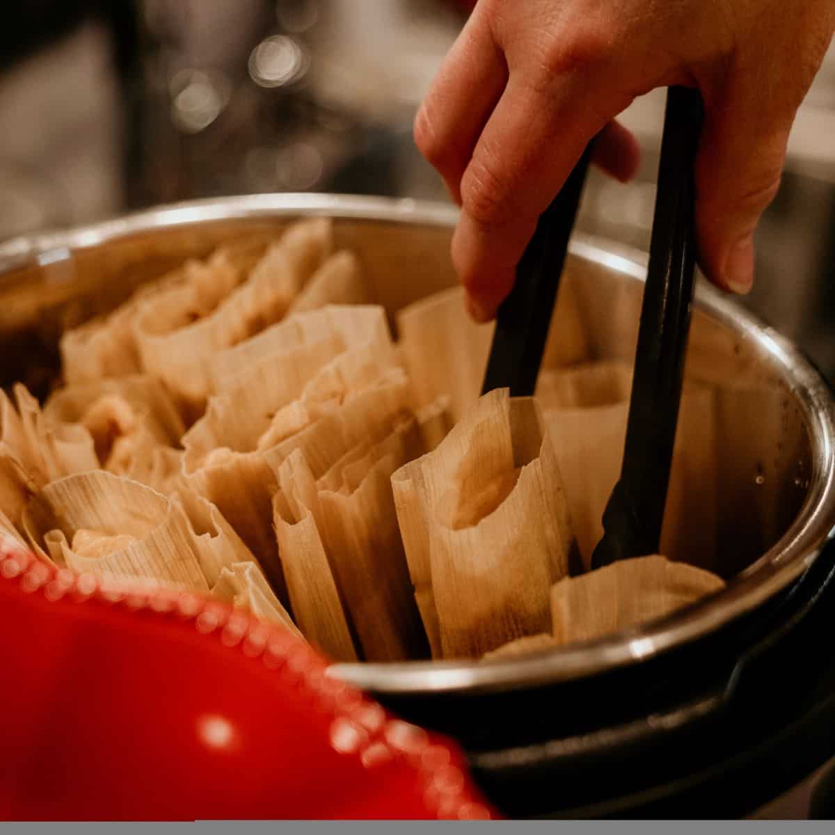 Tamales wrapped in dryed corn husk in a steamer basket with a red bowl in the foreground. A hand holding black tongs attempts to pull a single tamale from the steamer.