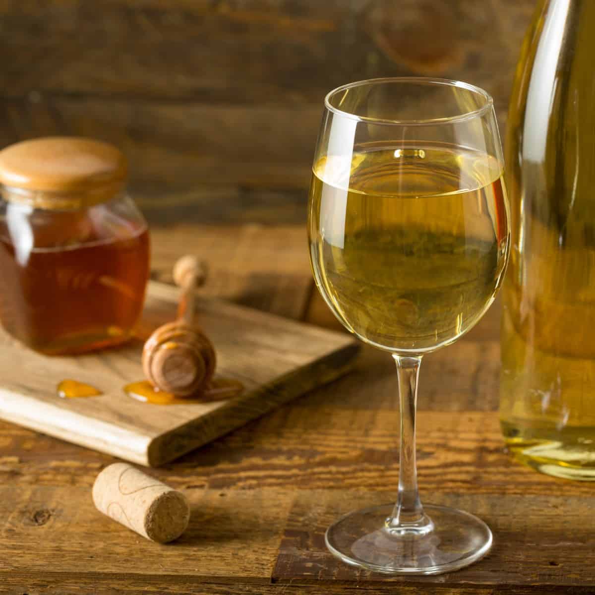 Wine glass with honey wine in forefront with a bottle of mead just behind it. To the left is a wood plank and cork with a jar of honey and honey dipper